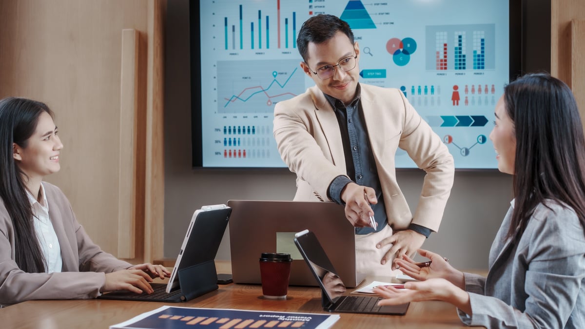 Business meeting in modern office: manager reviews data on laptop with two colleagues, analytics dashboard on screen, teamwork and strategy discussion.